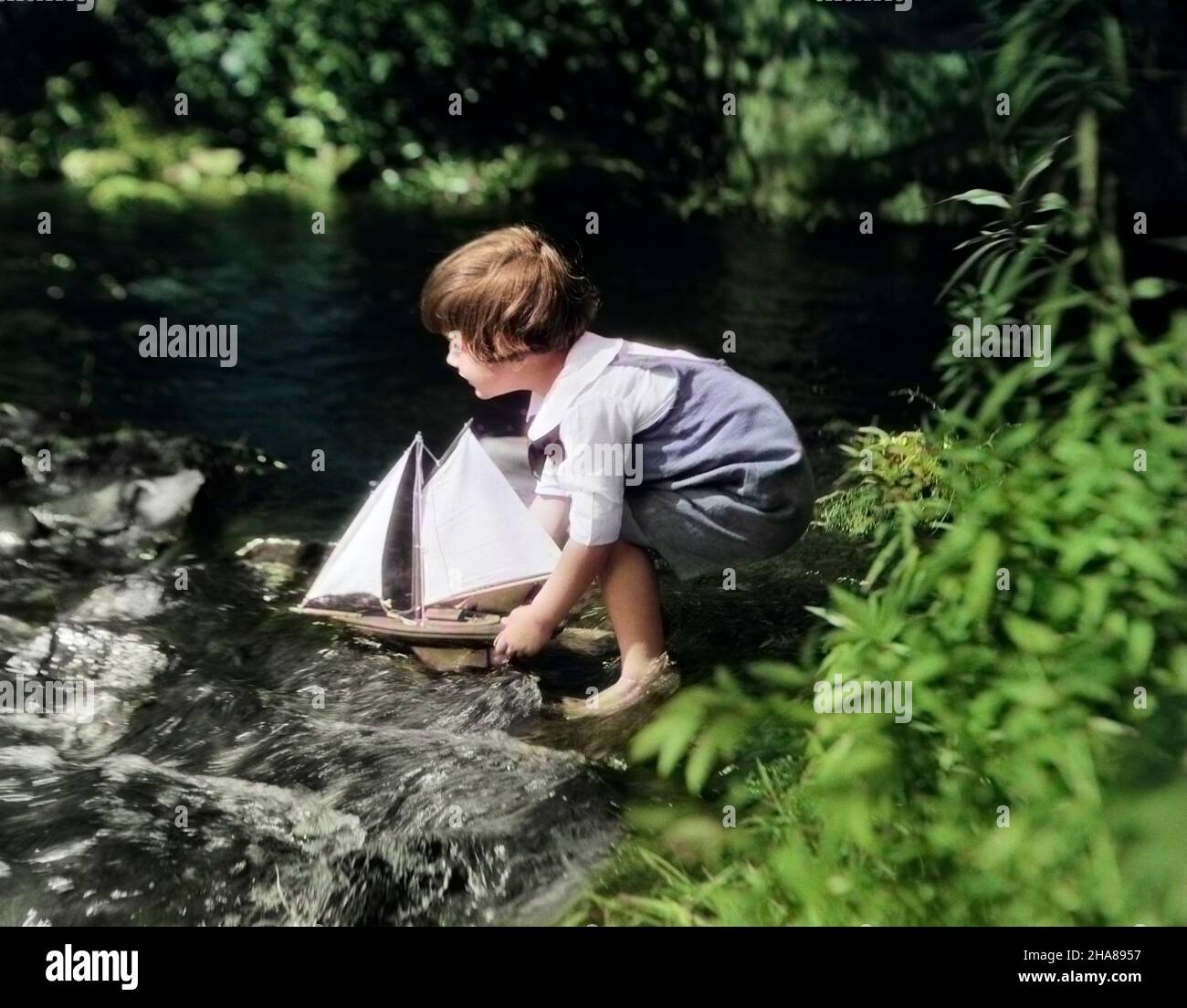 1920s LITTLE BOY PUTTING TOY SAILBOAT INTO STREAM - j128c HAR001 HARS TRANSPORTATION SUMMERTIME DREAMS HAPPINESS ADVENTURE RECREATION IMAGINATION MOBILITY PLEASANT AGREEABLE CHARMING JUVENILES LOVABLE PLEASING SEASON ADORABLE APPEALING CAUCASIAN ETHNICITY HAR001 OLD FASHIONED Stock Photo
