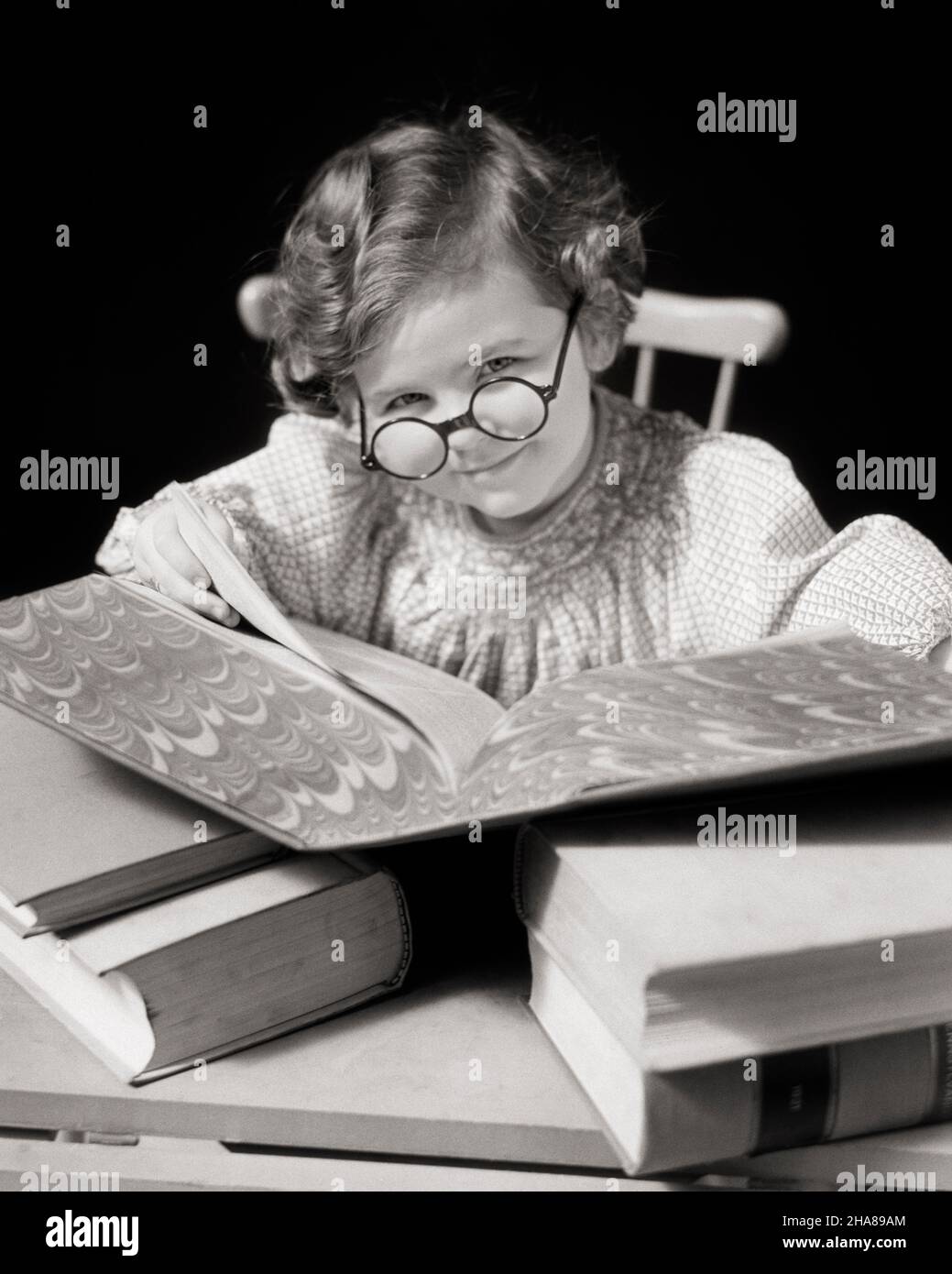 1930s LITTLE BRUNETTE GIRL SMILING READING USING A LARGE REFERENCE BOOK WEARING SCHOLARLY ROUND EYEGLASSES AND LOOKING AT CAMERA - j5098 HAR001 HARS HOME LIFE COPY SPACE CHARACTER CONFIDENCE EYEGLASSES B&W EYE CONTACT BRUNETTE SCHOOLS SUCCESS DREAMS GRADE REFERENCE HEAD AND SHOULDERS CHEERFUL DISCOVERY AND INTELLIGENT KNOWLEDGE PRIDE ACADEMIC OPPORTUNITY OCCUPATIONS PRIMARY SMILES USING JOYFUL PLEASANT SCHOLARLY SMART AGREEABLE CHARMING GRADE SCHOOL GROWTH JUVENILES LOVABLE PLEASING STUDIOUS ADORABLE APPEALING BLACK AND WHITE CAUCASIAN ETHNICITY HAR001 INQUISITIVE OLD FASHIONED Stock Photo