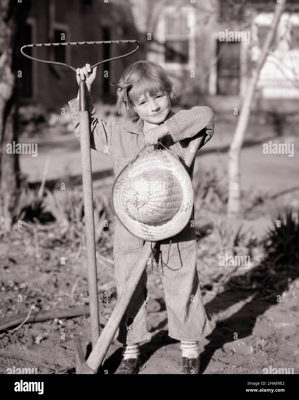 1930s CUTE LITTLE GIRL STANDING IN GARDEN LOOKING AT CAMERA WEARING HICKORY STRIPE COVERALLS HOLDING STRAW HAT PICK AX AND RAKE - j5764 HAR001 HARS PLEASED JOY LIFESTYLE PICK FEMALES RURAL RAKE HOME LIFE STRIPE COPY SPACE FULL-LENGTH INSPIRATION CARING CONFIDENCE AGRICULTURE B&W EYE CONTACT DREAMS HAPPINESS CHEERFUL ADVENTURE CHORE LEISURE STRENGTH AND CHOICE EXCITEMENT RECREATION PRIDE IN OPPORTUNITY SMILES CONCEPTUAL IMAGINATION JOYFUL STYLISH HICKORY AX CREATIVITY GROWTH IDEAS JUVENILES RELAXATION TASK BLACK AND WHITE CAUCASIAN ETHNICITY COVERALLS HAR001 OLD FASHIONED Stock Photo