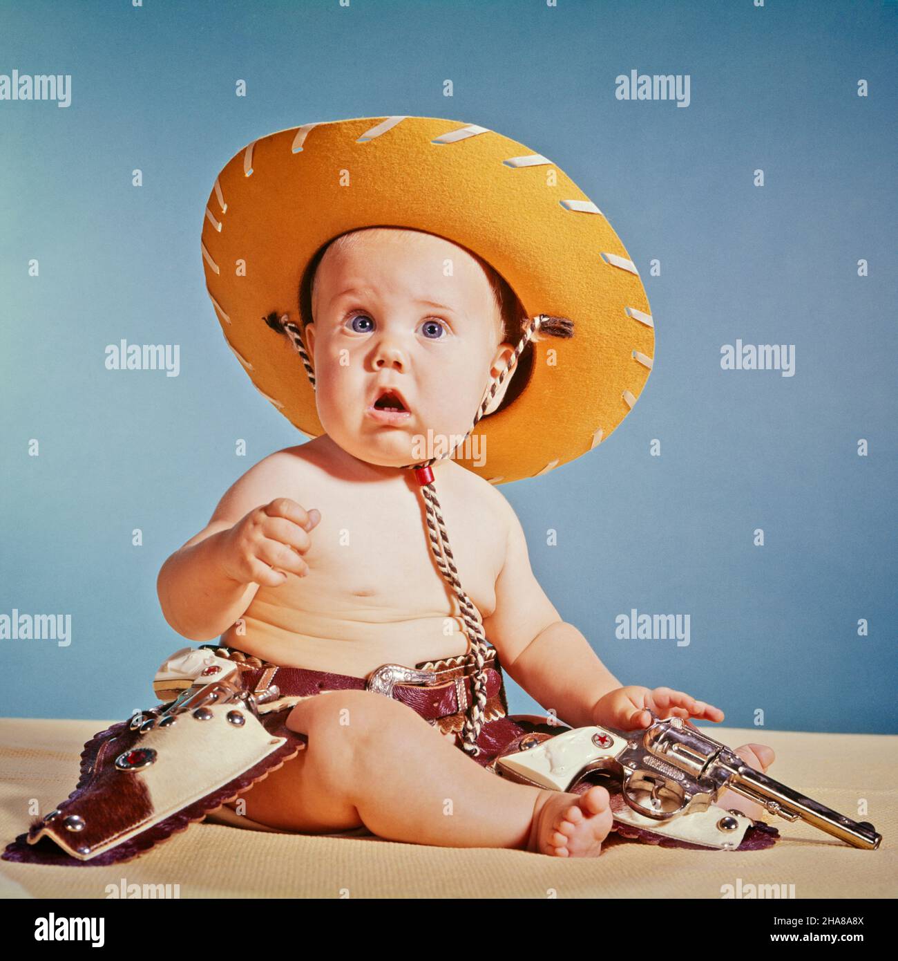 1960s BABY BOY WITH SURPRISED FACIAL EXPRESSION DRESSED AS COWBOY WEARING HAT AND HOLSTER AND GUN - kb6910 HAR001 HARS EYE CONTACT COWBOYS ADVENTURE EXCITEMENT BUNS SUPPORT HOLSTERS BABY BOY FIREARM FIREARMS JUVENILES CAUCASIAN ETHNICITY HAR001 OLD FASHIONED Stock Photo