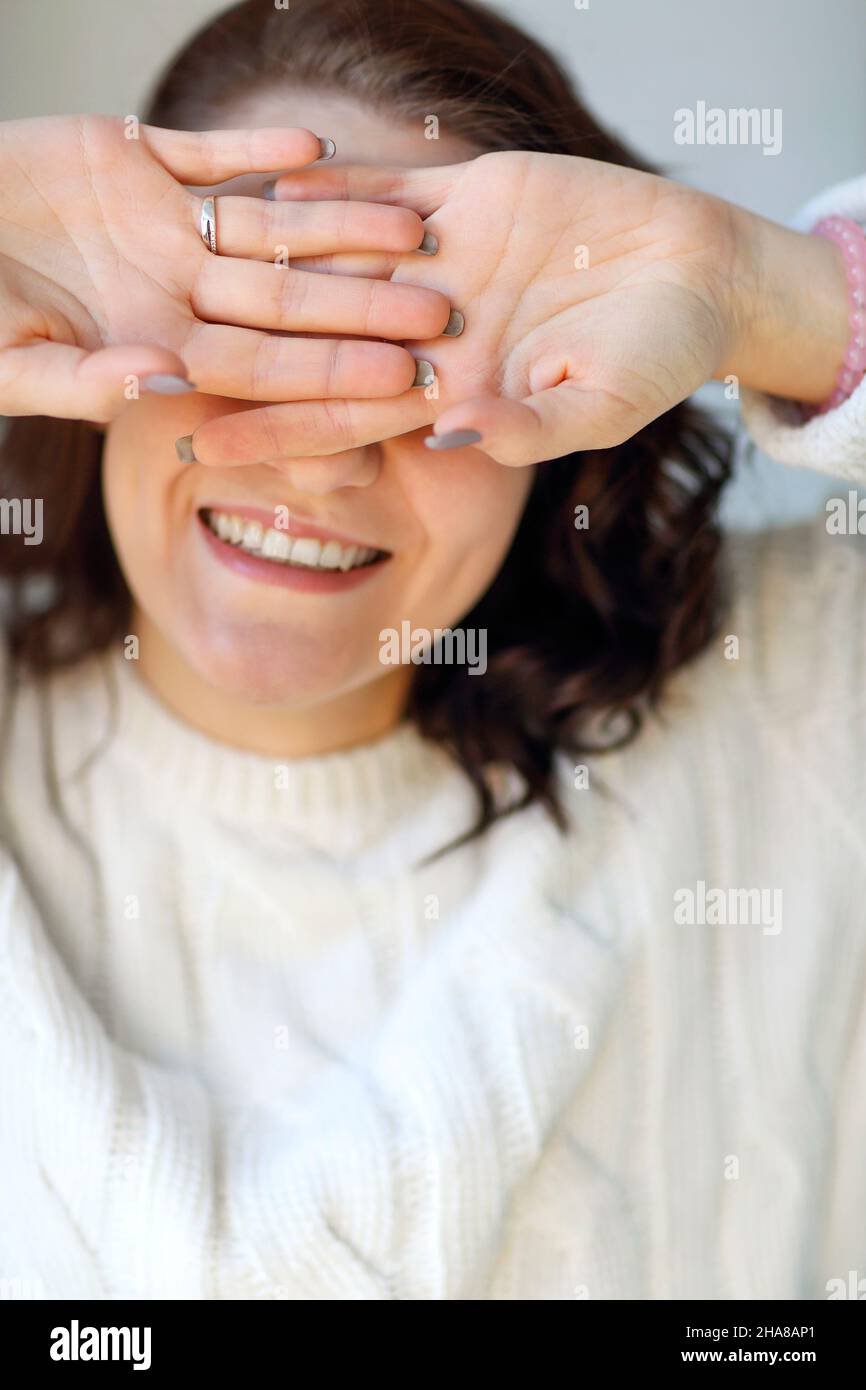 Happy delighted woman in knitted sweater closing her eyes with hands palms out and smiling on camera while spending leisure time at home, cheerful fem Stock Photo