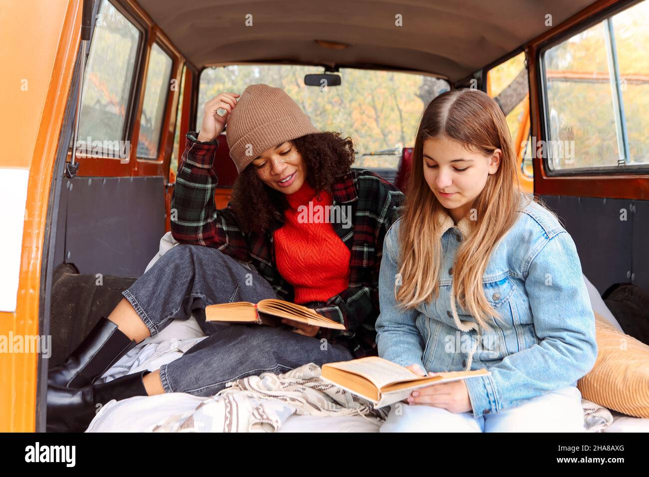 Two best interracial girlfriends sitting in wide open trunk of old vintage minivan car, couple of carefree happy students teenagers in warm clothes re Stock Photo