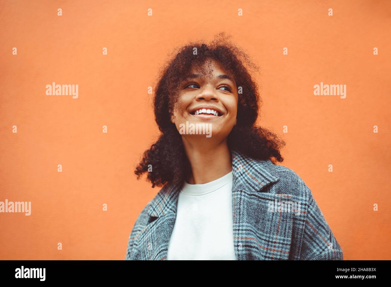 Enjoying Life. Beautiful happy African ethnicity girl with lush dark curly hairstyle in stylish coat standing against orange wall background, looking Stock Photo