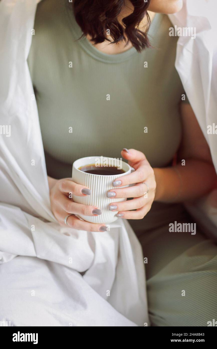 Relaxed positive young brunette woman in homewear sitting on top of unmade bed with cup of tea in her hands, slowly drinking aromatic hot beverage whi Stock Photo