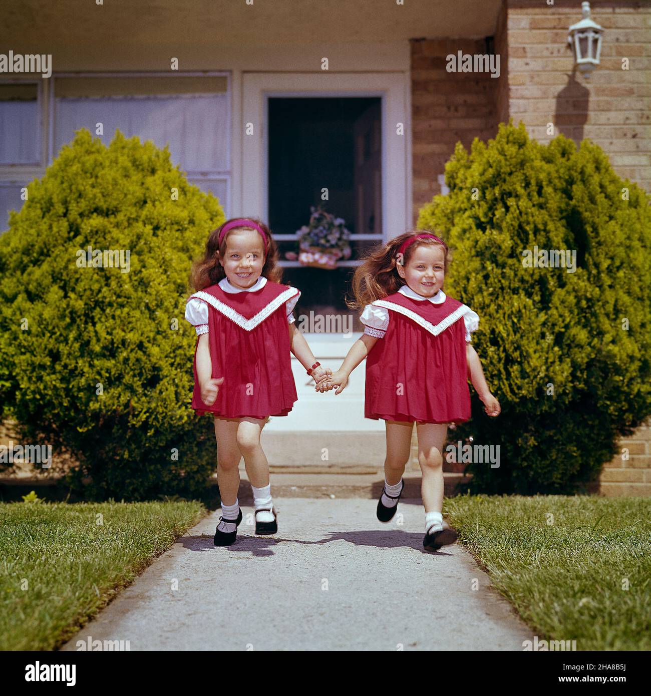 1960s TWIN GIRLS WEARING RED DRESSES MARY JANE SHOES RUNNING DOWN SIDEWALK FROM HOUSE HOLDING HANDS SMILING - kj3689 HAR001 HARS DOUBLE PLEASED JOY FEMALES MATCH CARING SIBLINGS SISTERS EXPRESSIONS DRESSES MATCHING SAME CHEERFUL MARY JANE HOLDING HANDS SIBLING SMILES JOYFUL LOOK-ALIKE PERSONAL ATTACHMENT AFFECTION DUPLICATE EMOTION JUVENILES LOOK ALIKE CAUCASIAN ETHNICITY CLONE HAR001 OLD FASHIONED Stock Photo