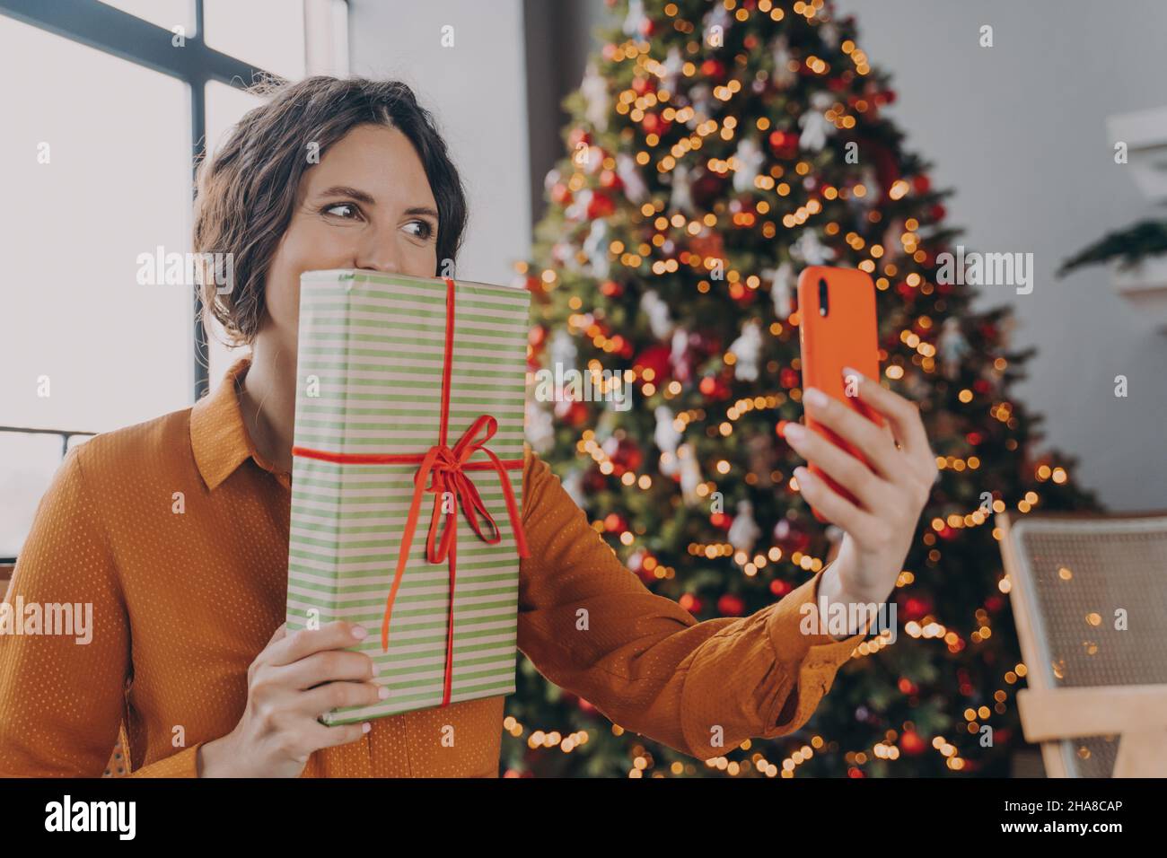 Joyful Spanish bright brunette lady making selfie on smartphone while posing with Xmas gift Stock Photo