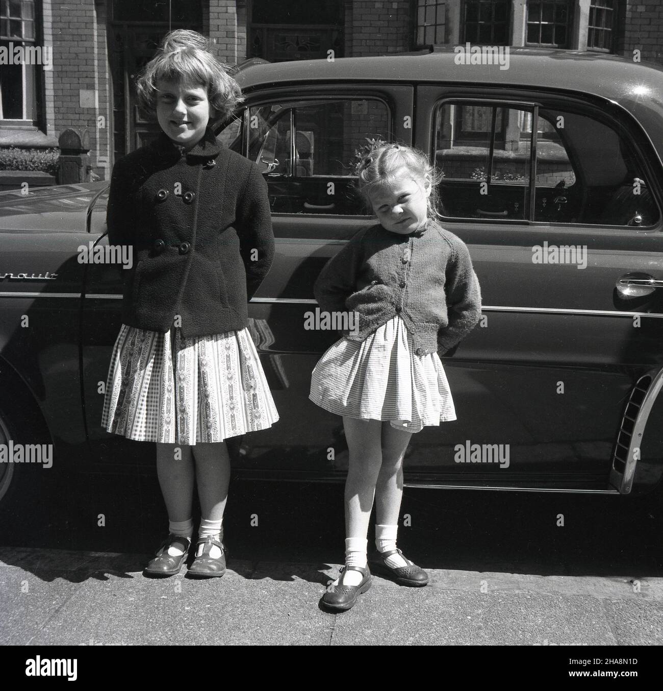 1958, historical, two young girls, sisters, stand outside in a British street by their father's car, a French Renault Dauphine, a rear-engined 4-door economy car, a successor to the Renault 4CV.  Between 1957 and 1961, Dauphines were assembled at Renault's Acton plant in West London and one was even driven HM the Queen. Roomy, with distinctive styling, the Dauphine car was a global success for Renault. Stock Photo