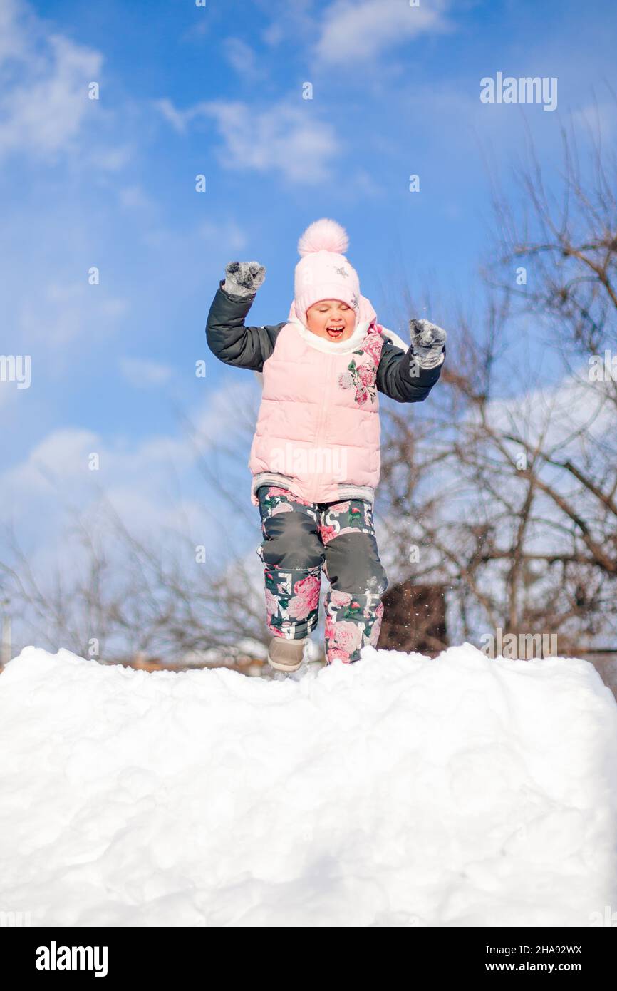 Little girl on top of snowdrift. Child in warm winter suit has fun, plays with snow and rides snow slide during walk in nature on sunny winter day Stock Photo