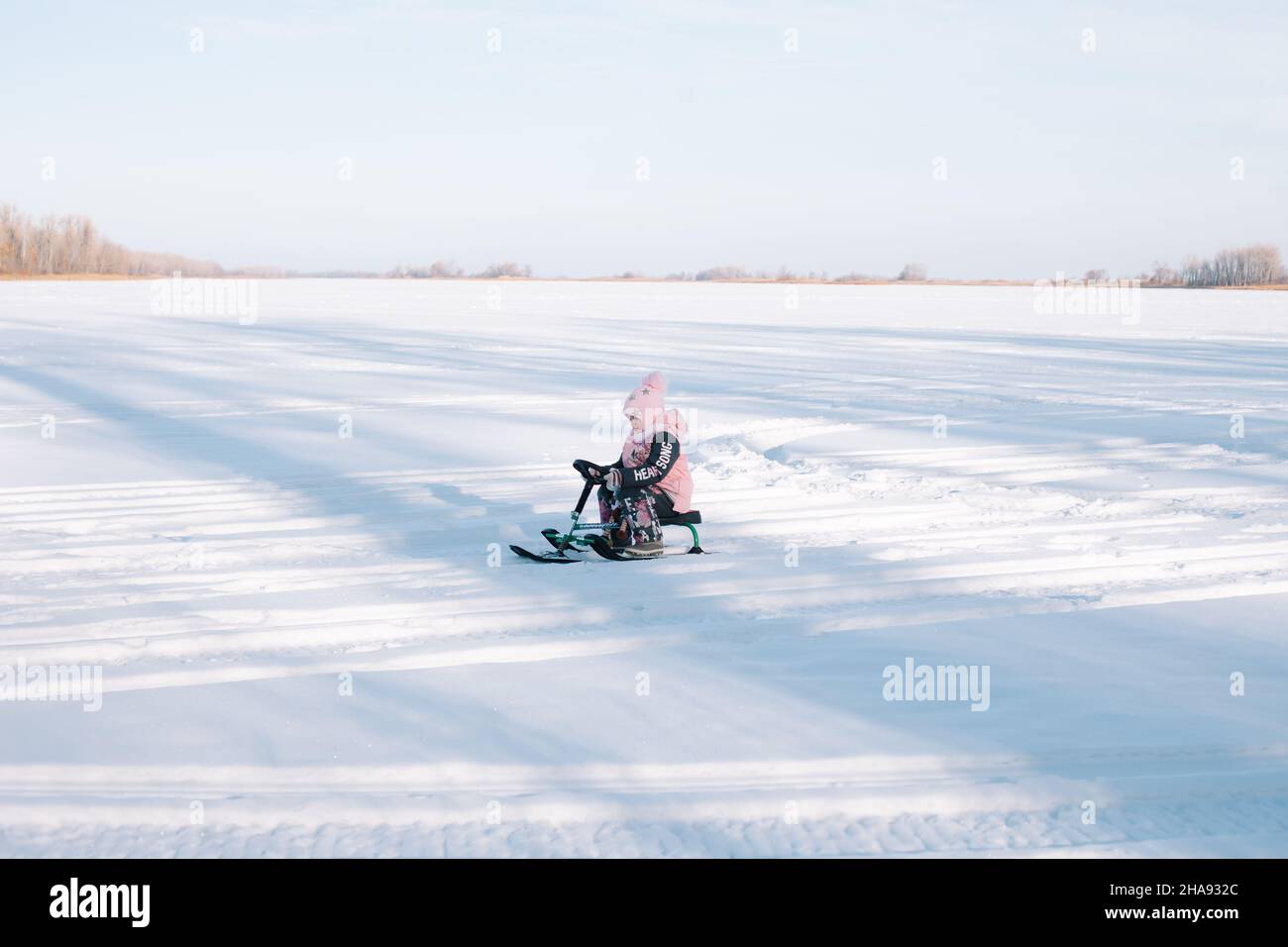 Child rides snowcat on snowy road. Little girl in pink warm jacket enjoys walk in nature and sledding on frozen river on sunny winter day, side view Stock Photo