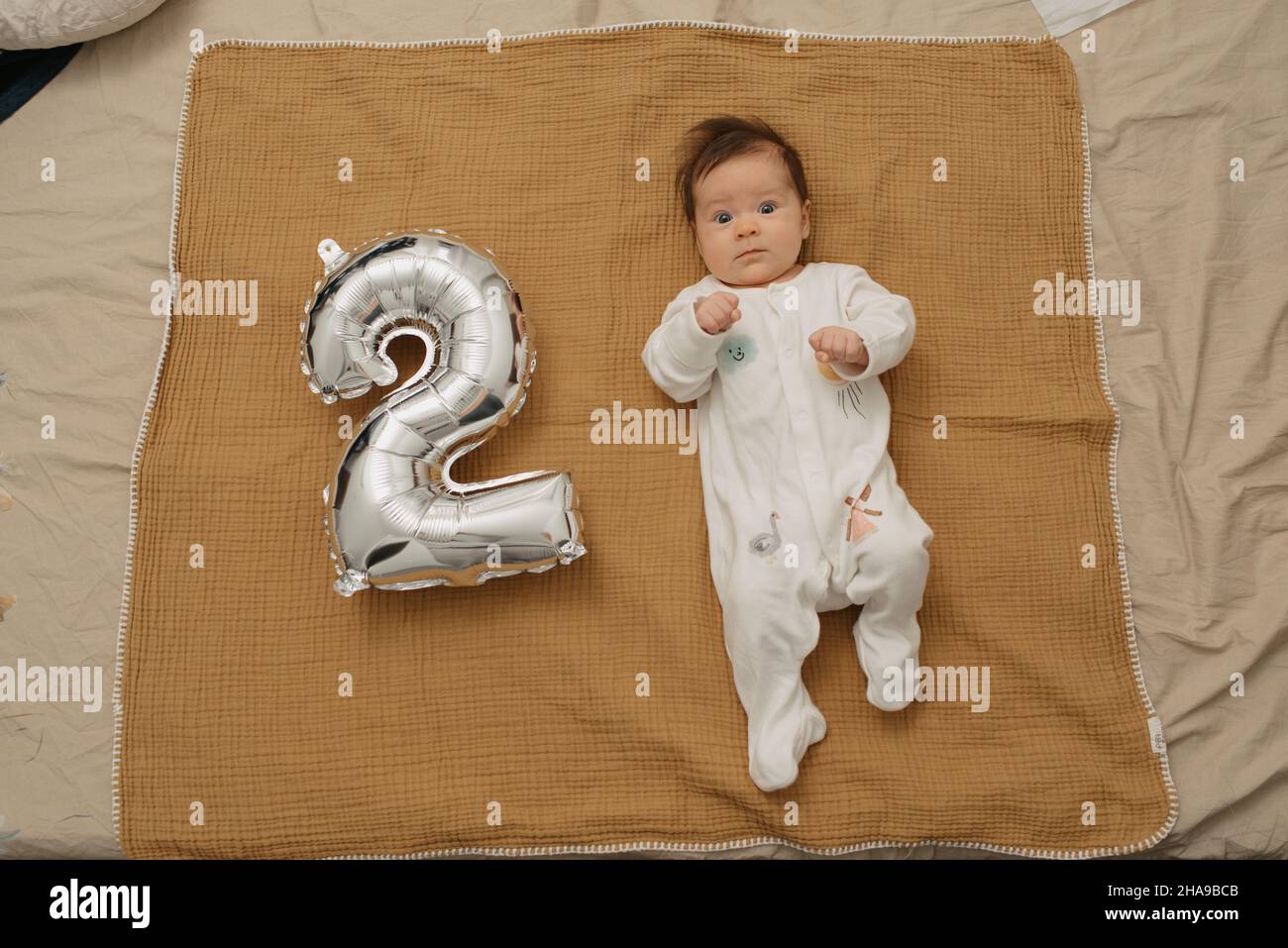 An Infant Is Lying On A Muslin Blanket Near A Silver Foil Balloon In The Shape Of 2 A Serious Baby Girl In One Piece Clothing Is Celebrating Her Two Stock Photo An Infant Is Lying On A Muslin Blanket Near A Silver Foil Balloon In The Shape Of 2 A Serious Baby Girl In One Piece Clothing Is Celebrating Her Two Stock Photo