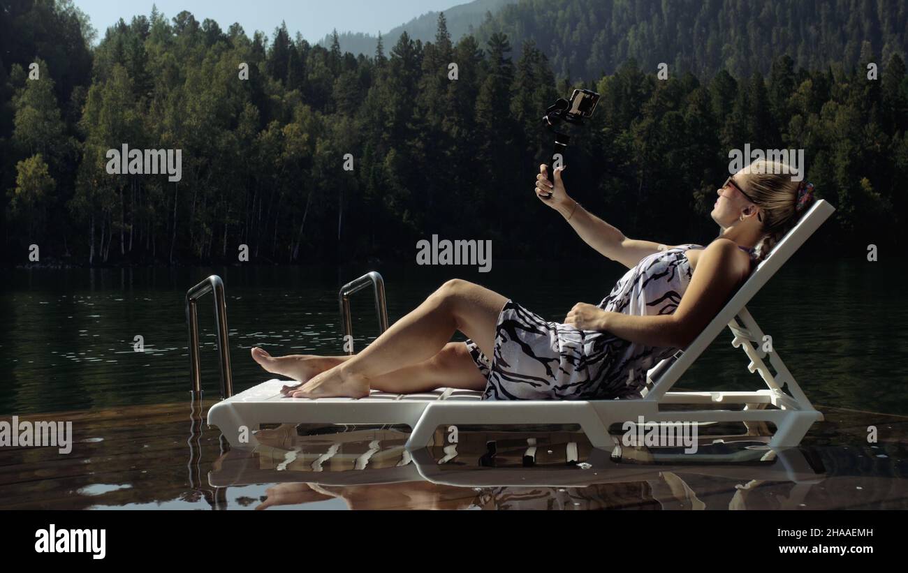 Woman shooting on handheld film gimbal stabilization for smartphone. Girl rest, relax, lie sunbed on pier in sunglasses, make selfie. Lady blogger bro Stock Photo