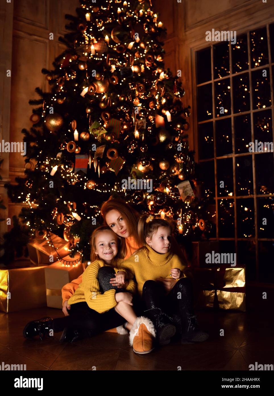 Happy mother and two small girls daughters sitting on floor together, hugging and looking at camera over decorated Christmas tree Stock Photo