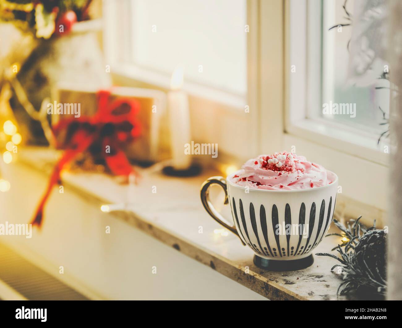 Mug with hot chocolate and whipped cream on window sill with candle, present and warm light. Hygge winter at home with warming drink. Stock Photo