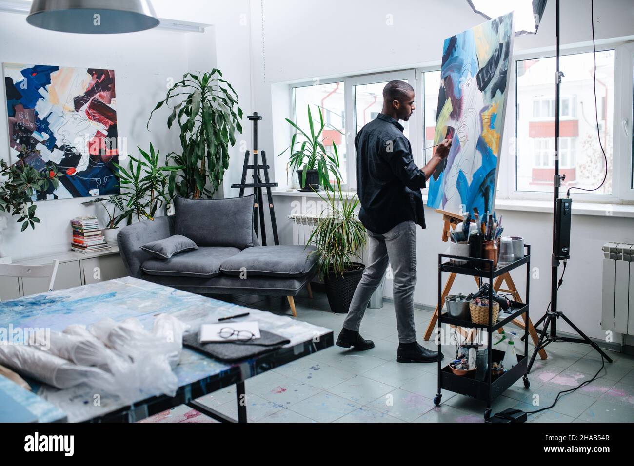 Talanted black man painting on an easel inside of his workshop in apartment. A sofa and an abstract painting on the wall behind. Window view on the bu Stock Photo