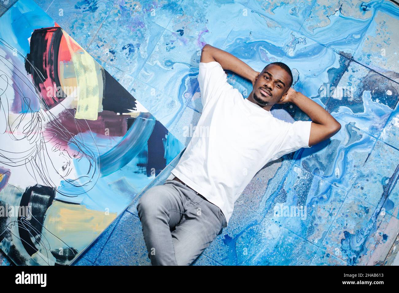 Black male artist lying on blue paint stained floor next to an abstract painting. Looking at the