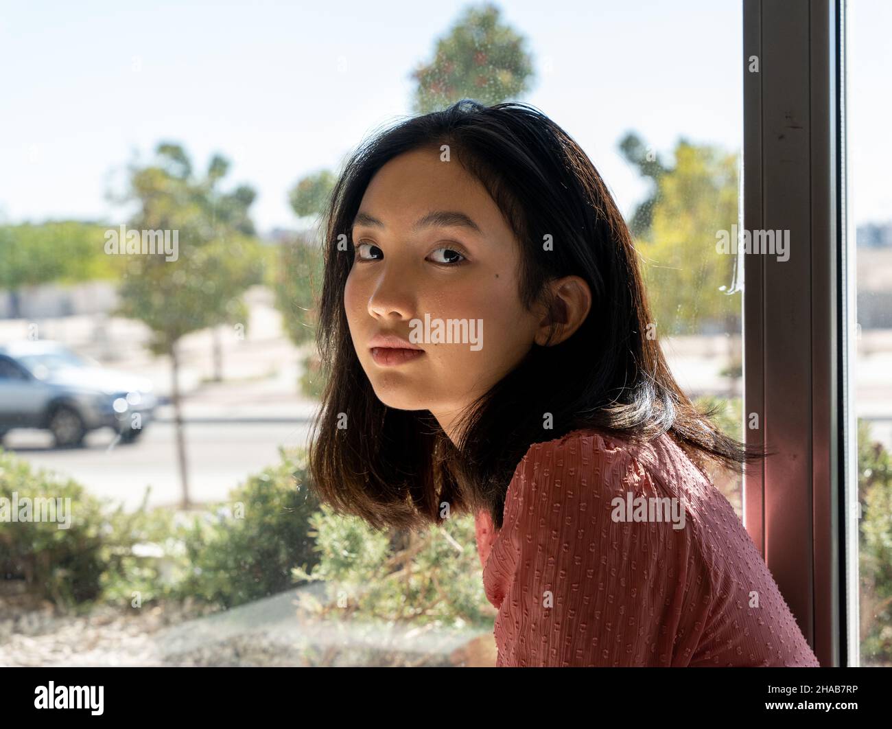 portrait of an Asian woman looking at the camera next to the window, horizontal urban background Stock Photo