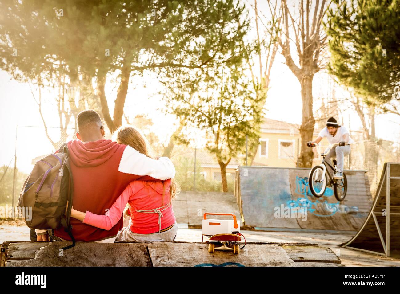 Multiracial couple in love sitting at skate park with music watching friends on bmx freestyle exhibition - Urban relationship concept Stock Photo