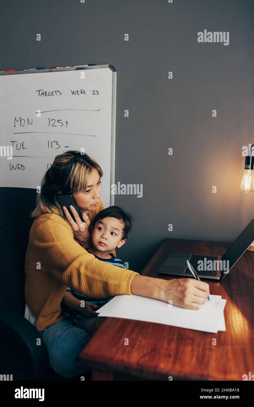Busy mom writing notes during a business phone call. Working mom carrying her son on her lap while sitting in her home office. Mother of one making bu Stock Photo
