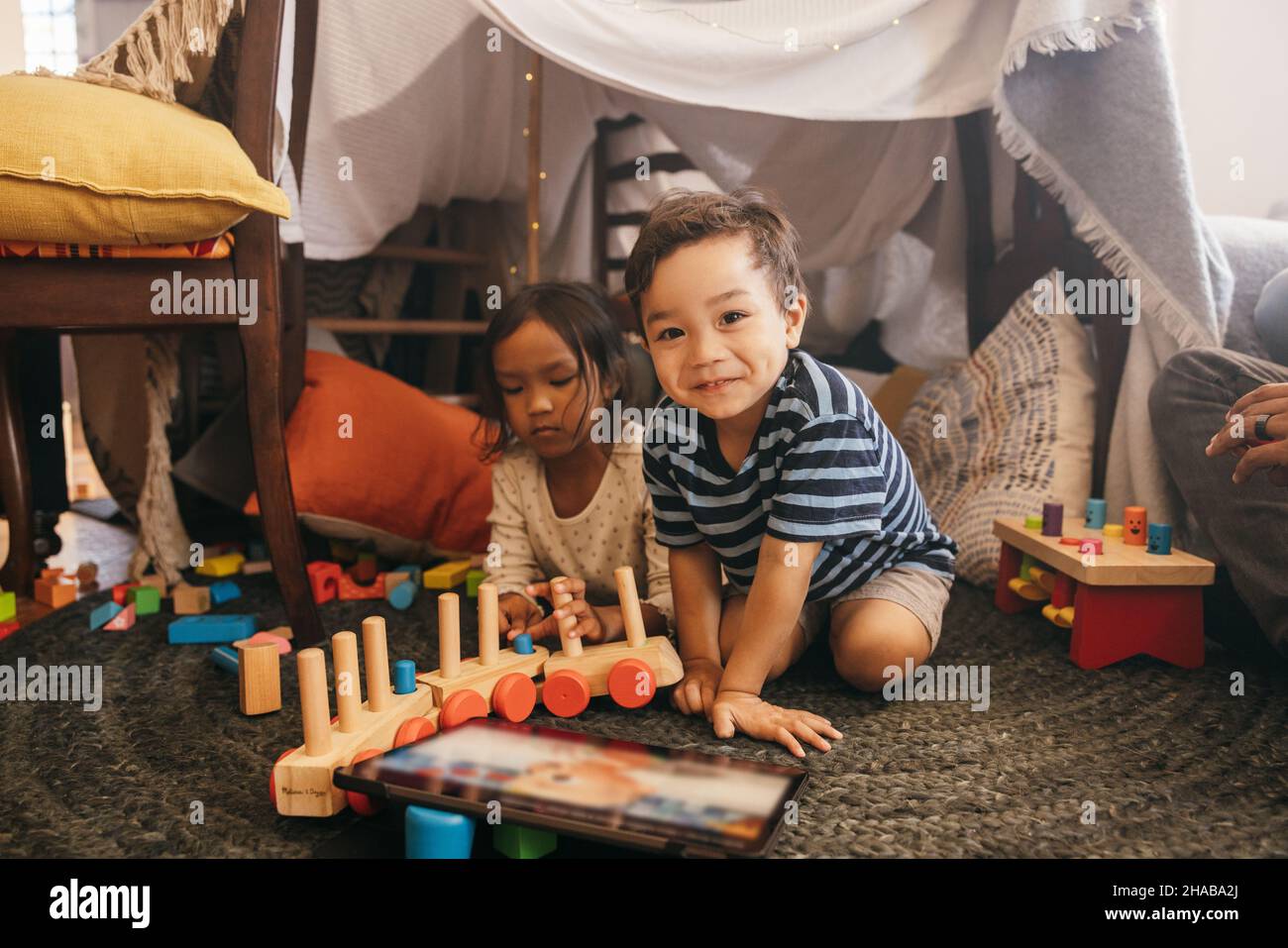 Cheerful little boy playing with his sister at home. Two adorable toddlers having fun in their play area with their parents sitting close by. Stock Photo