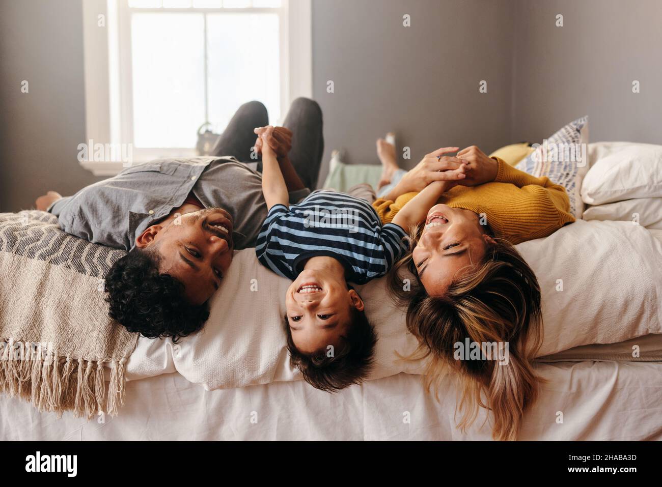 Happy family laughing and having fun together in their bedroom. Two ethnic parents playing with their son on the weekend. Mom and dad spending some qu Stock Photo