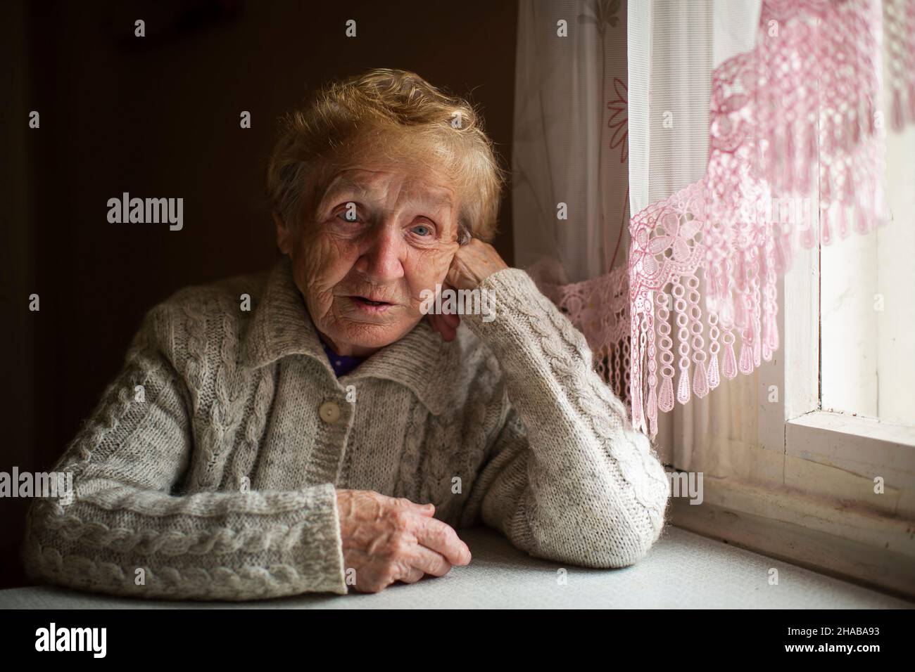 A distraught and puzzled old woman sits by the window in her house ...