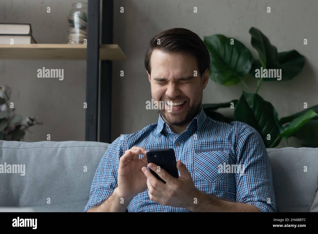 Man sits on sofa holds smartphone laughs on funny video Stock Photo