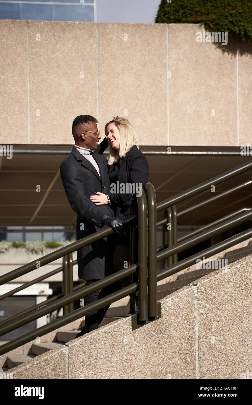 Happy black man and blond woman embracing each other and smiling while standing on stairway on modern city street in daytime Stock Photo