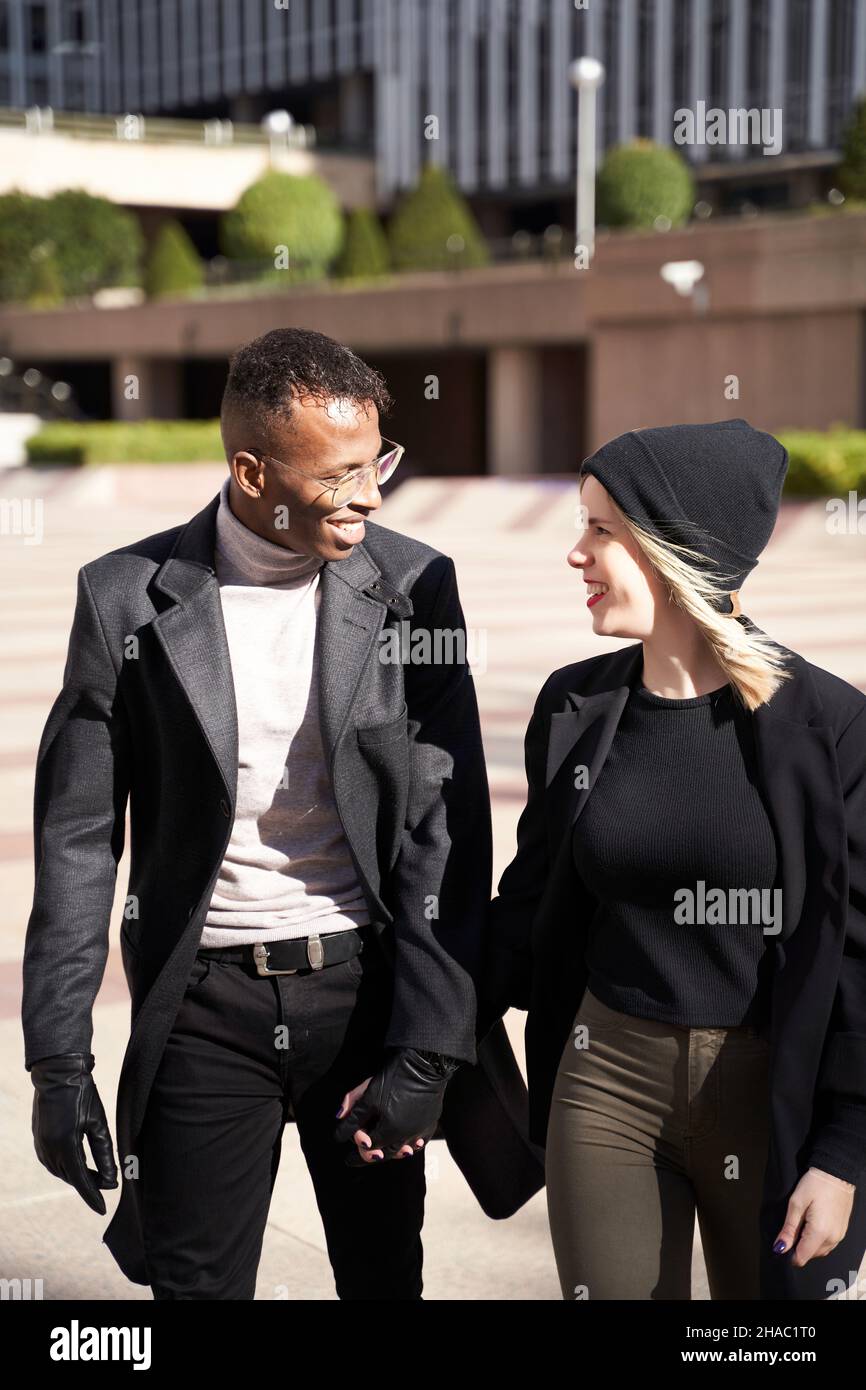 Happy multiethnic man and woman in casual clothes holding hands and looking at each other while strolling on city street during romantic date, on sunny day Stock Photo