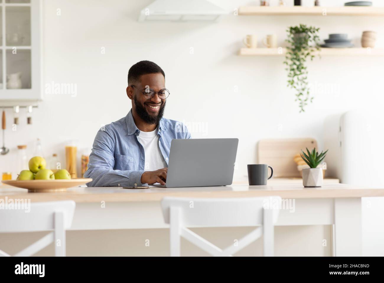 Smiling millennial african american bearded guy in glasses working on laptop in minimalist kitchen interior Stock Photo