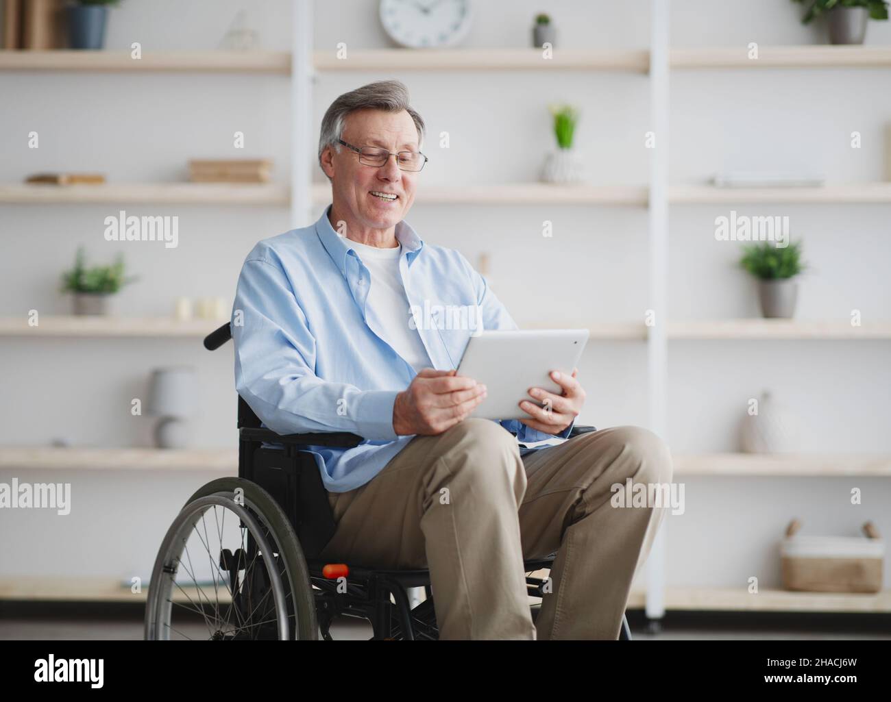 Cheerful senior man in wheelchair using tablet computer, browsing internet at home Stock Photo