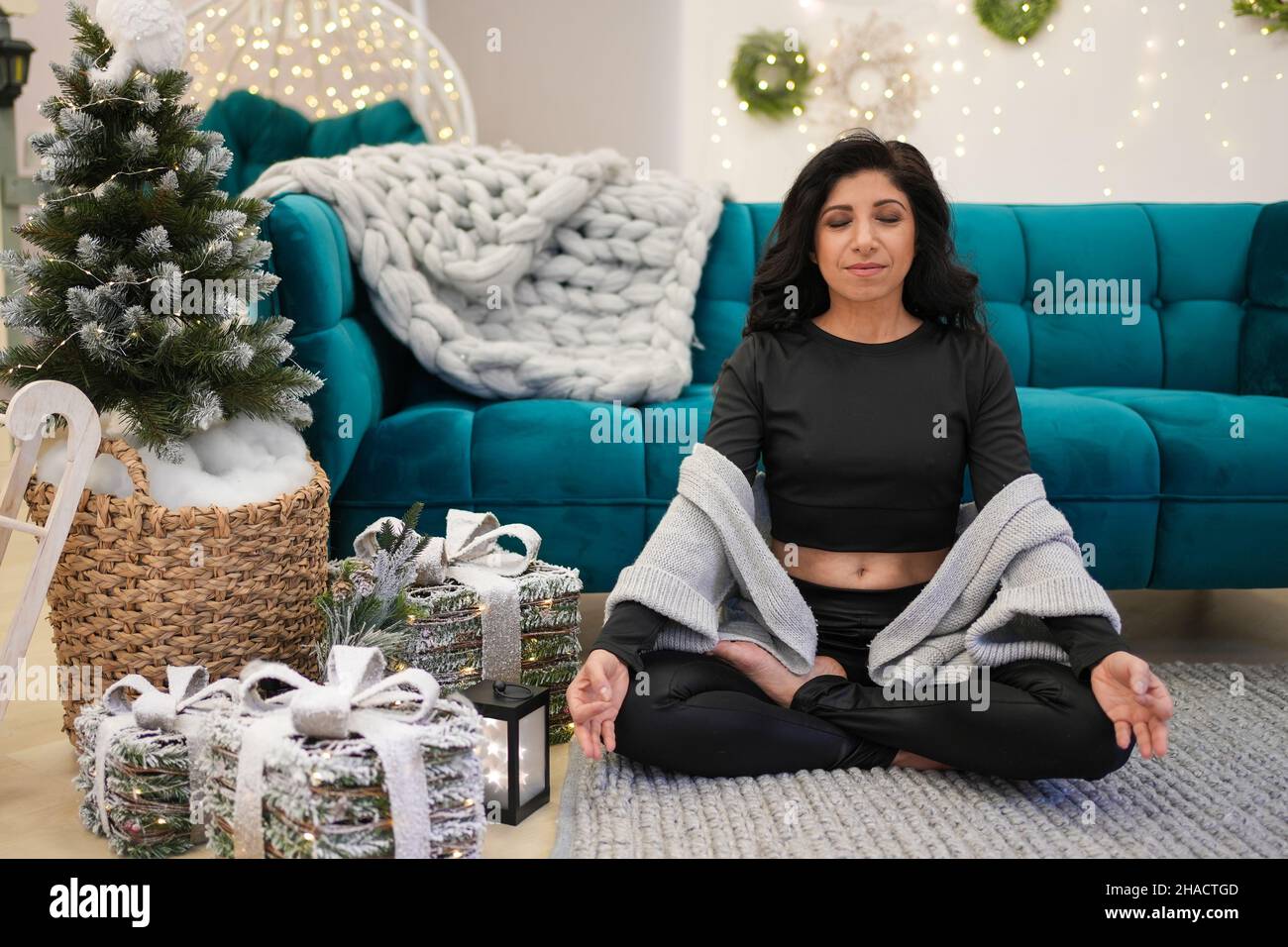 Portrait of middle age woman meditating while sitting near sofa and Christmas tree Stock Photo