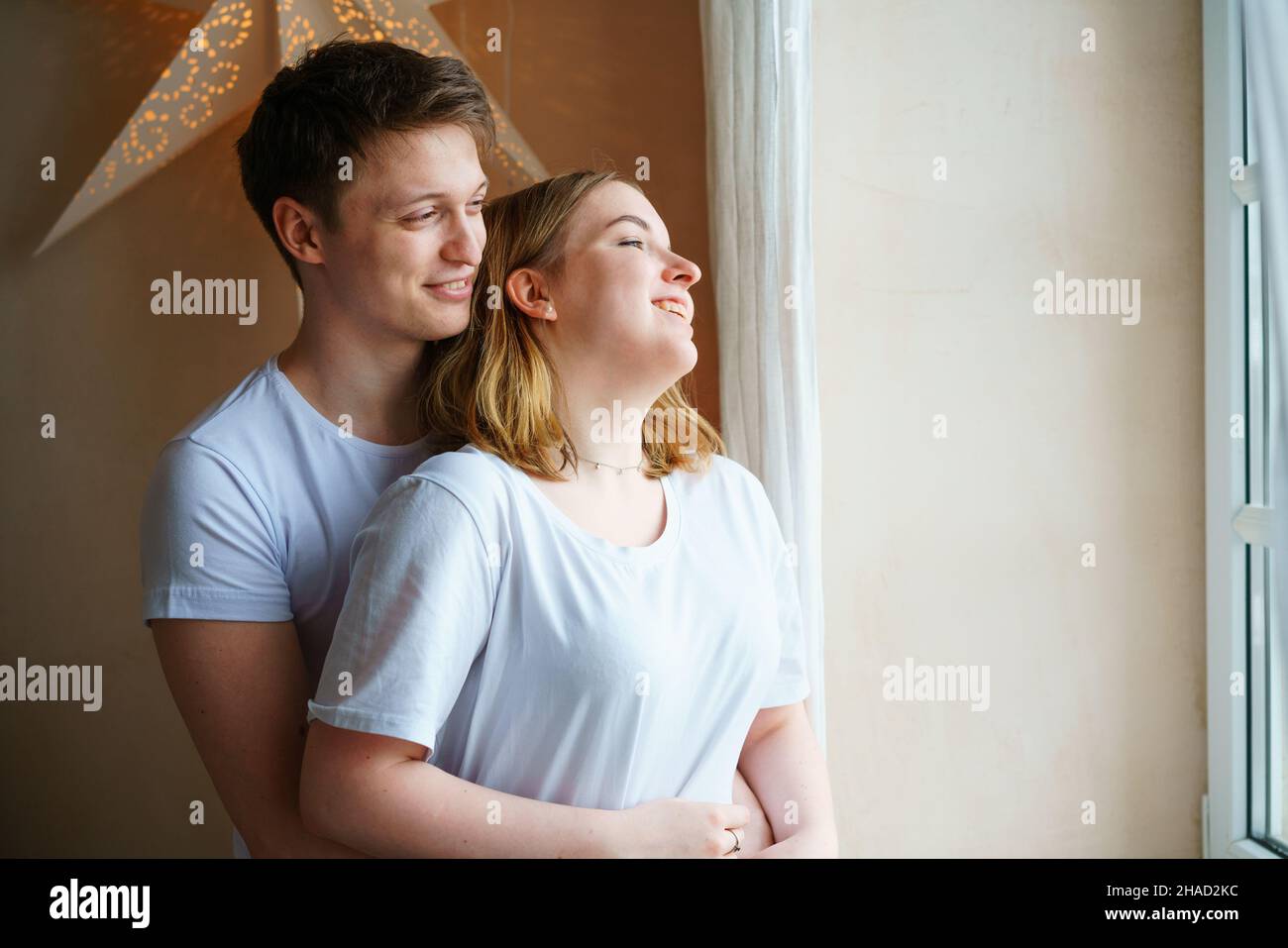 Shot of happy young couple standing together by window while looking ...