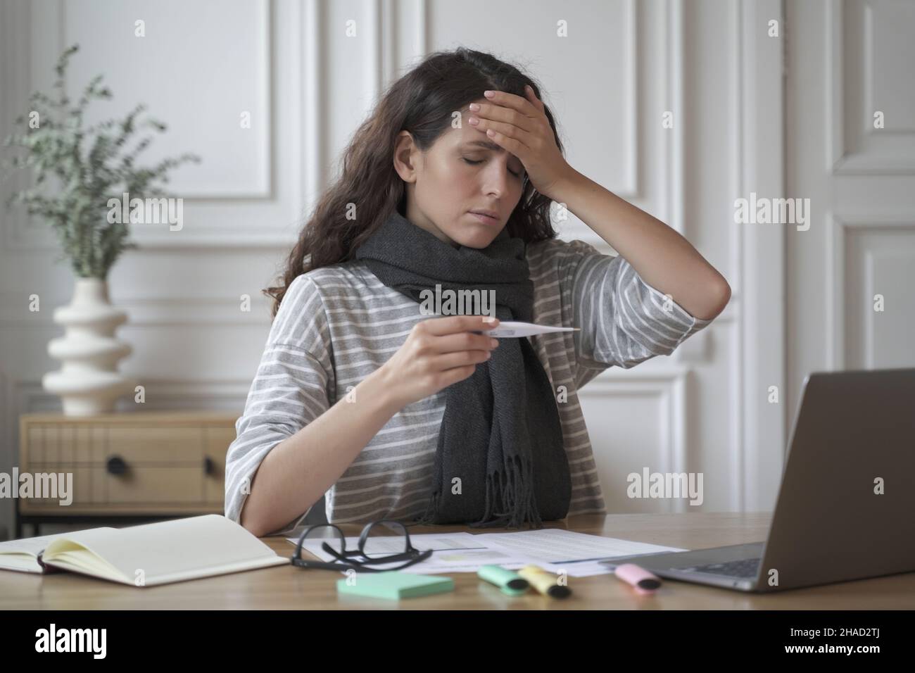 Sick young woman freelancer sitting at workplace and touching forehead while taking body temperature Stock Photo