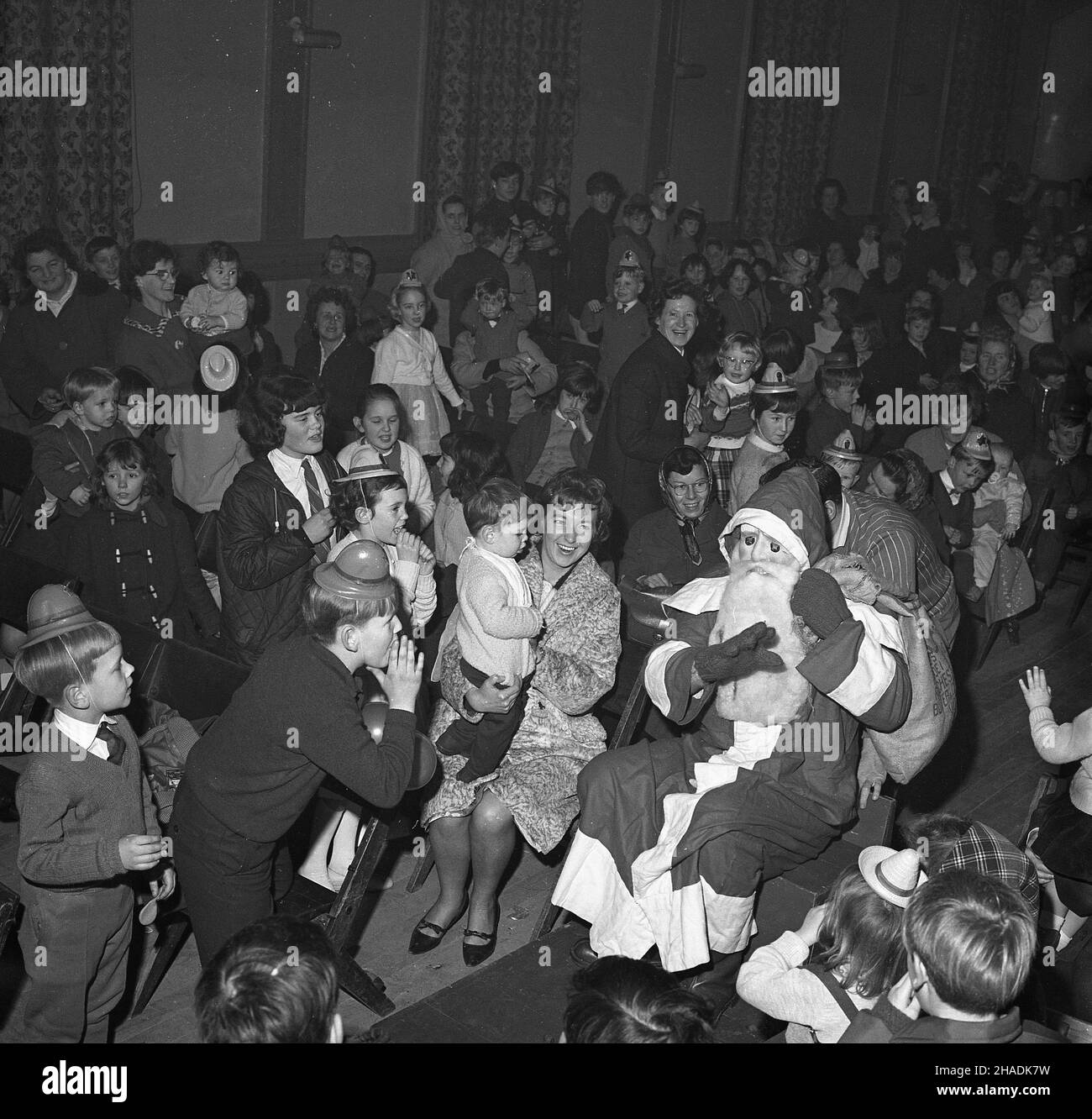 1965, historical, a santa claus in costume, mask and beard, with sack of presents, sitting on his wooden sledge which is being pushed into the middle of a village hall to the delight of the young children, who have just had a christmas party. Stock Photo