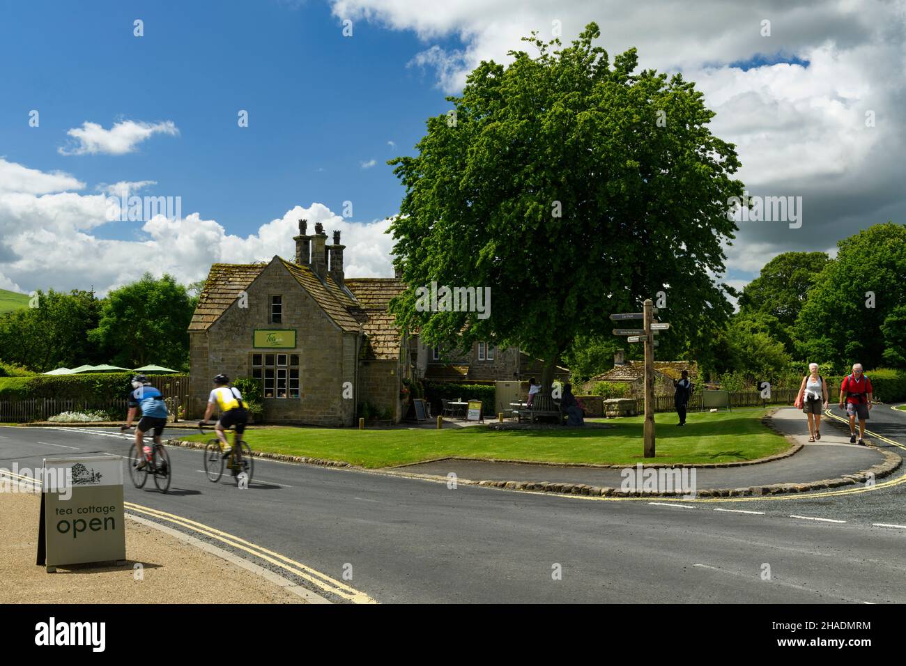 2 cyclists & walkers passing quaint attractive cottage tea rooms cafe in scenic sunny rural village - B6160 Bolton Abbey, Yorkshire Dales, England UK. Stock Photo