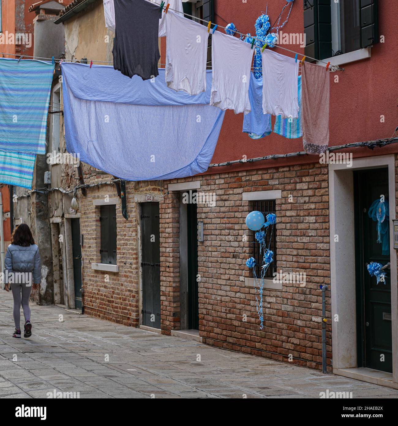 A young woman walks through a narrow alley in Venice. Laundry hangs to dry on linen above her. Blue party balloons hang from windows and doors. Stock Photo