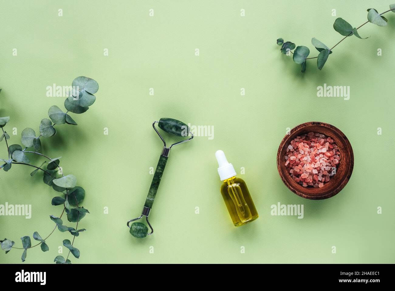 Cosmetic serum in glass bottle, pink himalayan salt, face roller and eucalyptus leaves on light green background. Skincare, natural organic cosmetics Stock Photo