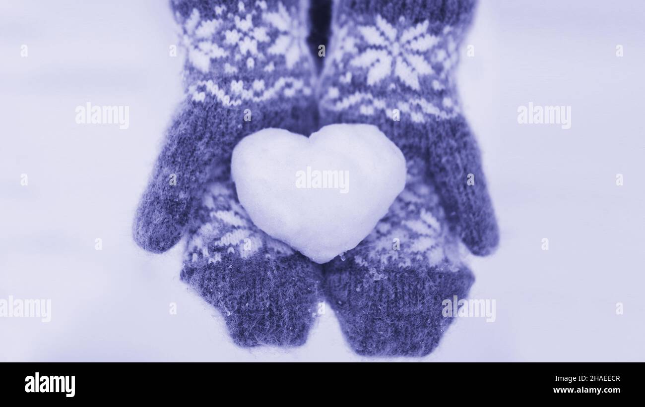 Color of the Year 2022 Very Peri. A girl in knitted red mittens holding a heart made of snow on her palms. Valentine's day concept. Declaration of Stock Photo