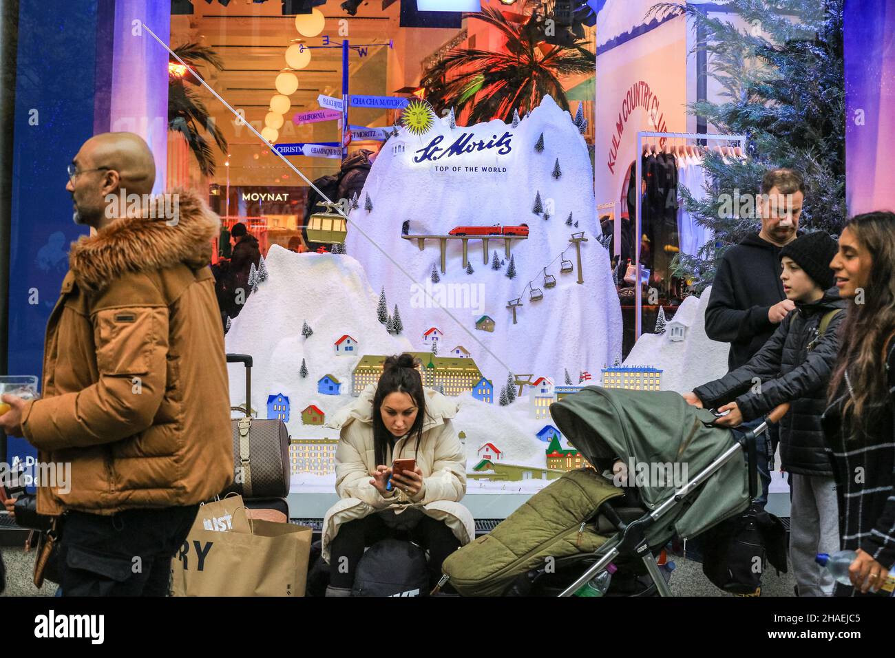 Oxford Street London, UK, 12th Dec 2021. A family take a break from shopping outside a window decorated with a snowy mountain scene. Shoppers and visitors to London crowd into busy Oxford Street today despite infection rates with the new omnicron covid variant in London, and the wider country, rising fast, and government considering the introduction of further measures to curb the trend. Stock Photo
