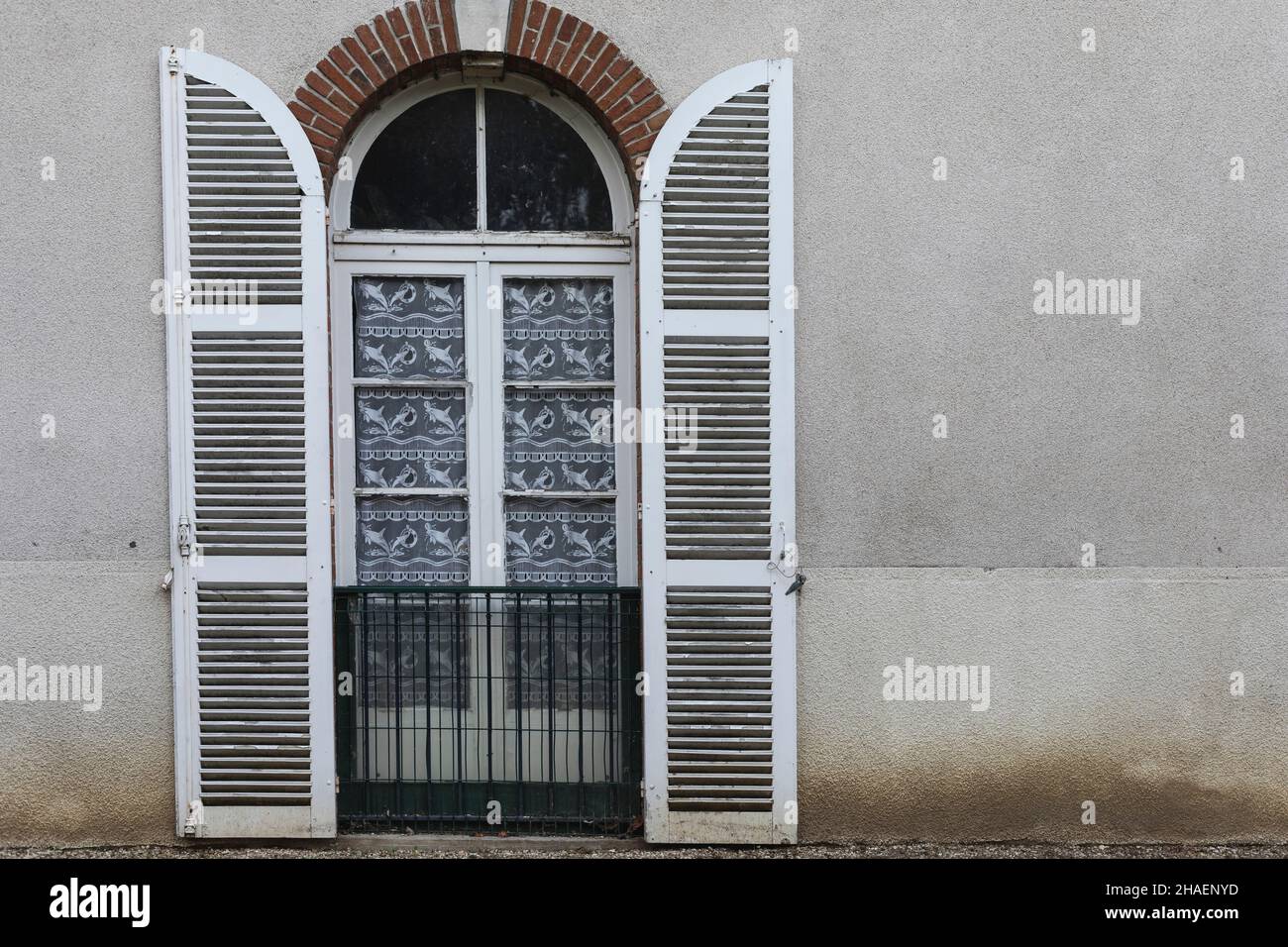 white painted plantation shutters open and pinned back against a wall to reveal a glass window with white net curtains Stock Photo