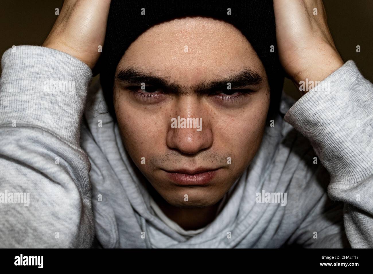 young Filipino man close up of face with hands on head, angry concentrating face. white sweater black hat bushy eyebrows red eyes Stock Photo