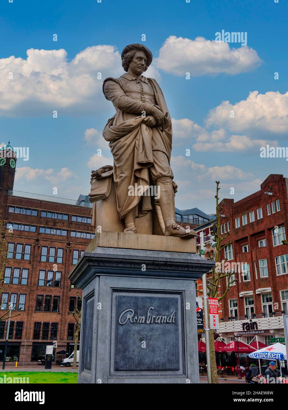 Amsterdam, Netherlands - May, 2021: Closeup of bust of Rembrandt statue under blue cloudscape, with clock tower in back on Rembrandtplein. Stock Photo