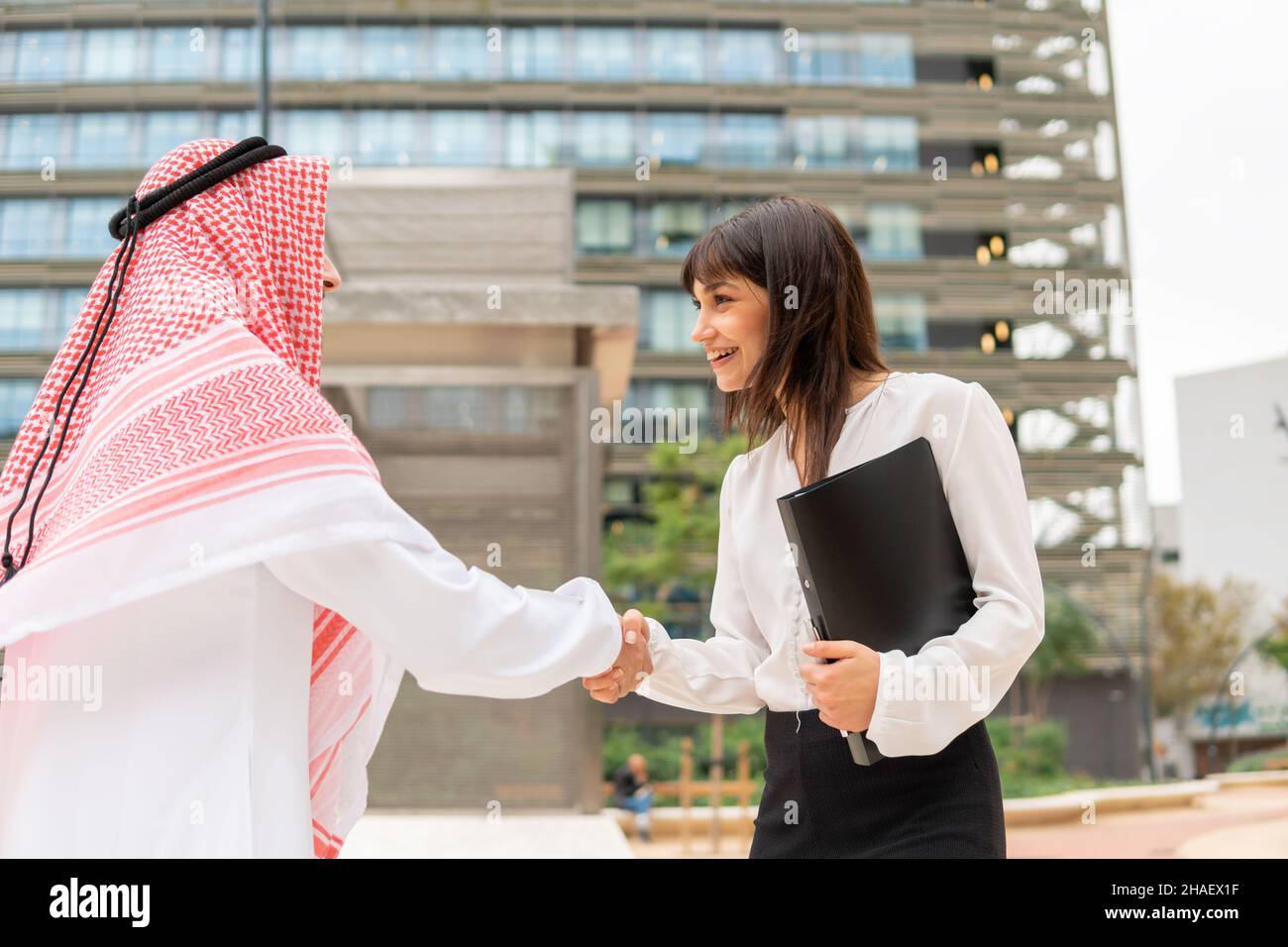Young pleasant caucasian businesswoman greeting arab male client at meeting outdoors, two smiling business partners saudi man and european woman shaking hands after making successful deal Stock Photo