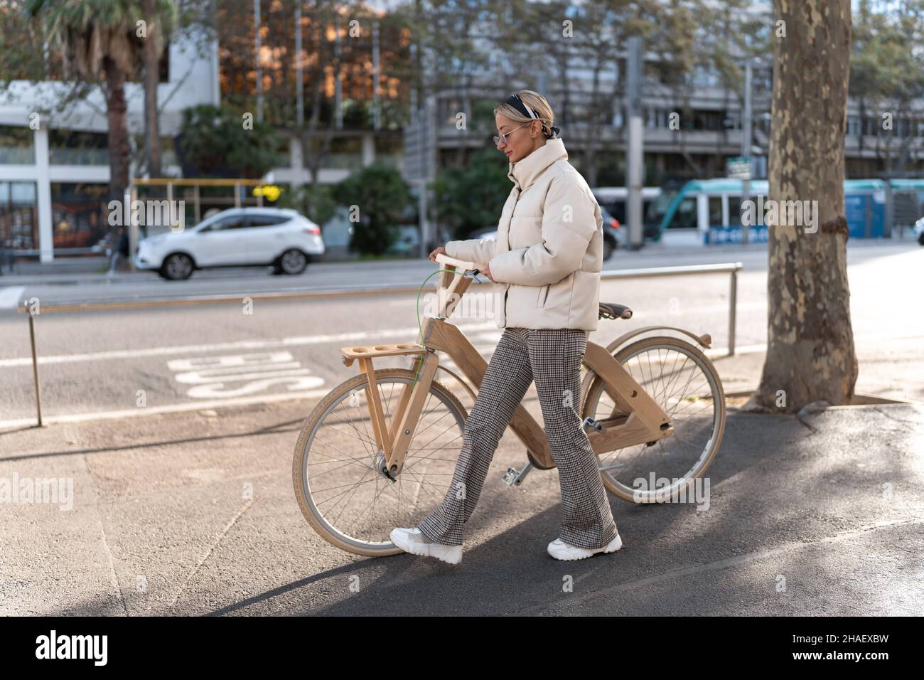 Full body woman in outerwear pushing eco friendly bike while walking on sidewalk near road on sunny day in city. Stock Photo
