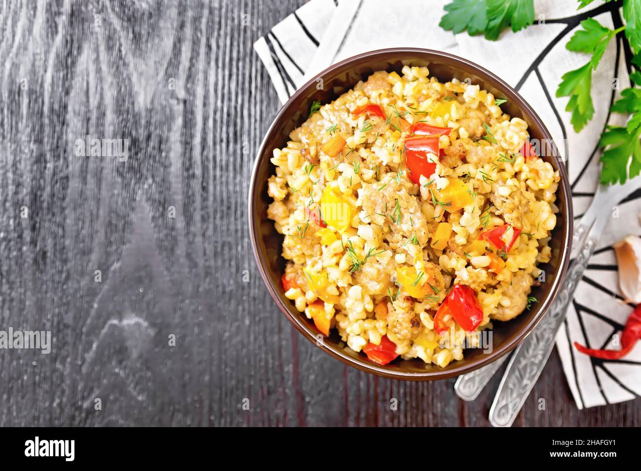 Barley porridge with minced meat, yellow and red bell peppers, garlic and onions in a clay bowl, napkin and parsley on wooden board background from ab Stock Photo