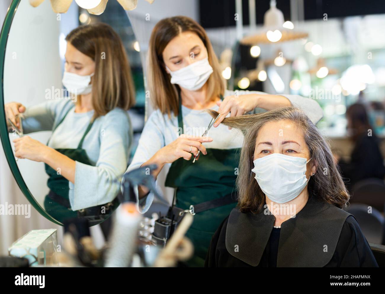 Skillful woman hair stylist wearing face mask making haircut to aged