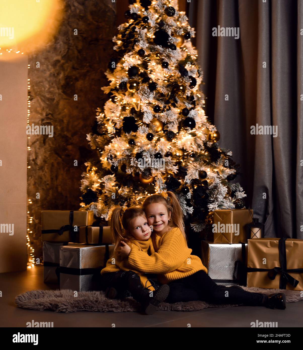 Happy small girls sisters in yellow sweaters sitting on floor together, hugging looking at camera over decorated Christmas tree Stock Photo