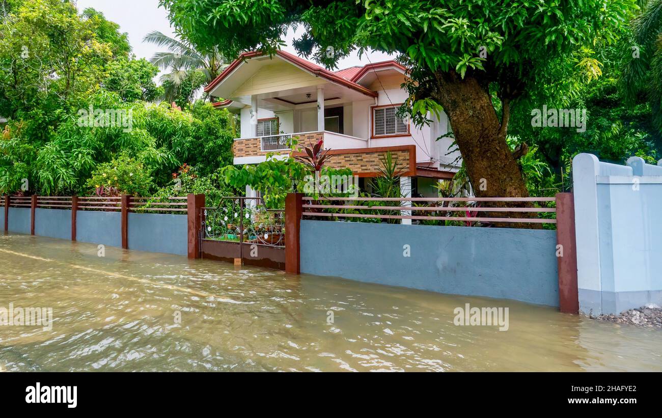 Heavy monsoon rains cause water on a flooded highway to rise up the front wall and entrance gate outside the yard of a two-story home in Baco, Orienta Stock Photo