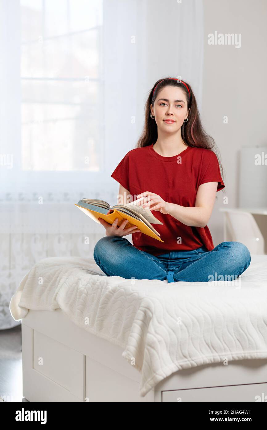 A young Caucasian woman is sitting cross-legged on the bed and leafing through a book. In the background, the white interior of the room. The concept Stock Photo