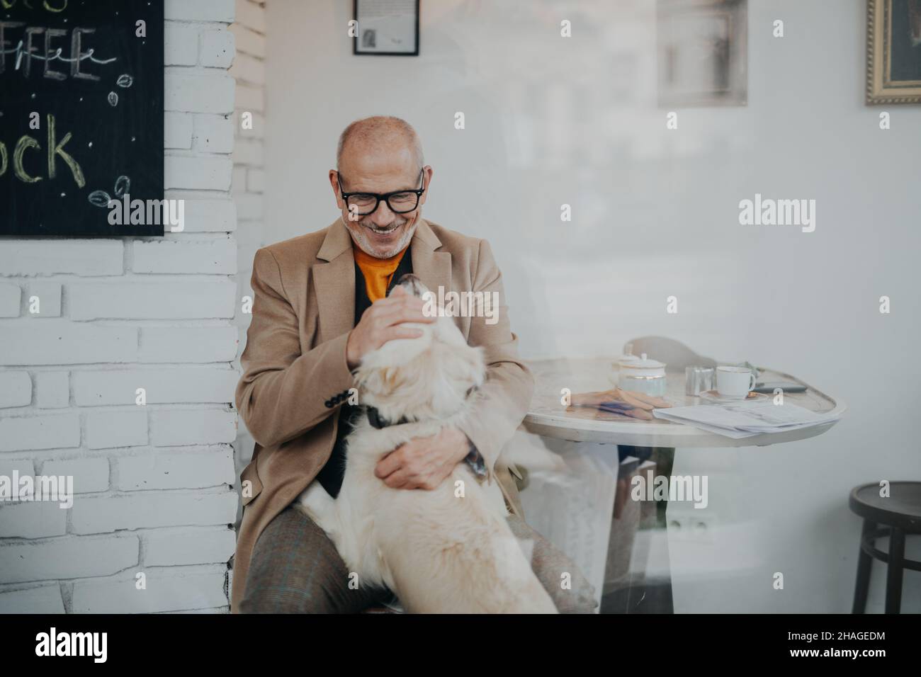 Happy senior man sitting in cafeteria and playing with his dog, shot through glass. Stock Photo